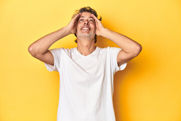 Middle-aged man posing on a yellow backdrop laughs joyfully keeping hands on head. Happiness concept.