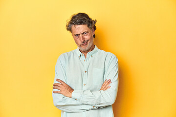 Middle-aged man posing on a yellow backdrop frowning face in displeasure, keeps arms folded.