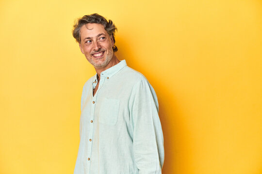 Middle-aged man posing on a yellow backdrop looks aside smiling, cheerful and pleasant.