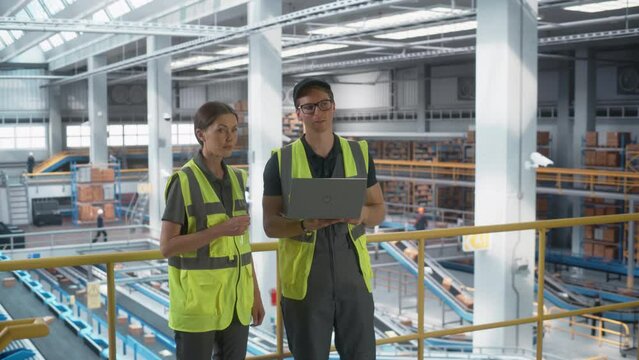 Sortation Fullfilment Center: Supervisor and Manager Talk, use Laptop. Logistics and Transportation Warehouse Facility with Conveyor Belt Loading Cardboard Boxes with Goods for Delivery to Customers