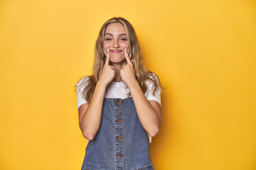 Young blonde Caucasian woman in denim overalls posing on a yellow background, doubting between two options.