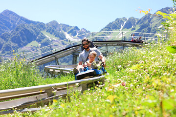 father and child having ride on summer toboggan called Rodelbahn rushing down the track . Beautiful mountains on background - obrazy, fototapety, plakaty