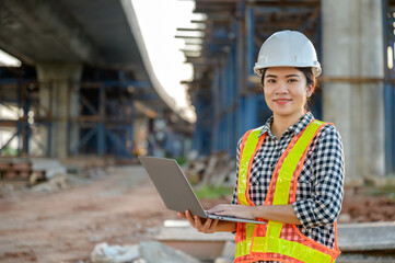 Portrait of an asian female road construction engineer or civil engineer using a laptop computer standing on a road construction site to supervise the construction of a new road