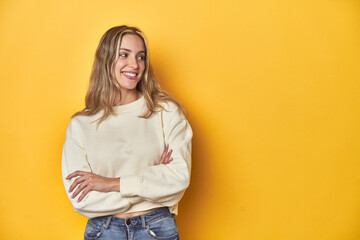 Young blonde Caucasian woman in a white sweatshirt on a yellow studio background, smiling confident...