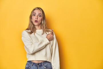 Young blonde Caucasian woman in a white sweatshirt on a yellow studio background, pointing to the...