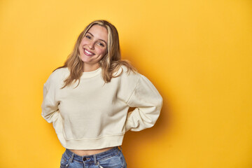 Young blonde Caucasian woman in a white sweatshirt on a yellow studio background, happy, smiling...