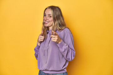Young blonde Caucasian woman in a violet sweatshirt on a yellow background, pointing to front with fingers.