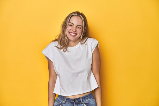 Young blonde Caucasian woman in a white t-shirt on a yellow studio background, laughs and closes eyes, feels relaxed and happy.