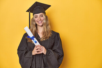 Young graduate woman in cap and gown, holding a diploma on a yellow studio background.