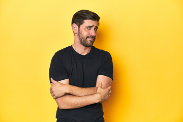 Caucasian man in black t-shirt, yellow studio backdrop suspicious, uncertain, examining you.