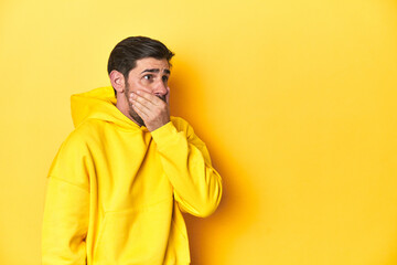 Man in yellow hoodie, monochrome studio backdrop thoughtful looking to a copy space covering mouth with hand.