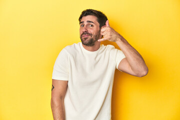 Caucasian man in white t-shirt on yellow studio background showing a mobile phone call gesture with fingers.