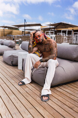 Chico joven tatuado y musculoso posando en terraza de chiringuito en vacaciones