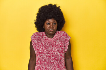 African-American woman with afro, studio yellow background shrugs shoulders and open eyes confused.