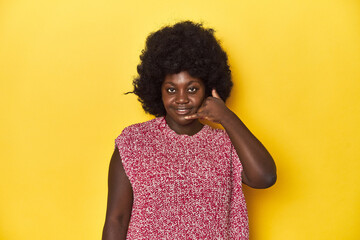 African-American woman with afro, studio yellow background showing a mobile phone call gesture with fingers.