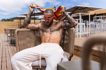 Chico joven tatuado y musculoso posando en terraza de chiringuito en vacaciones