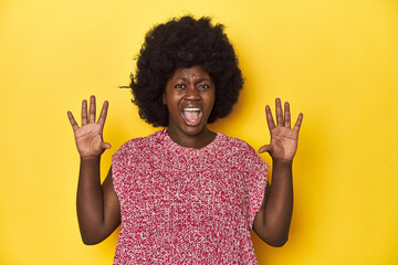 African-American woman with afro, studio yellow background receiving a pleasant surprise, excited and raising hands.