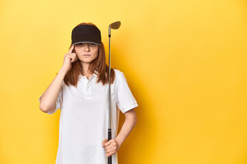 Redhead golfer with club and cap, studio shot pointing temple with finger, thinking, focused on a task.