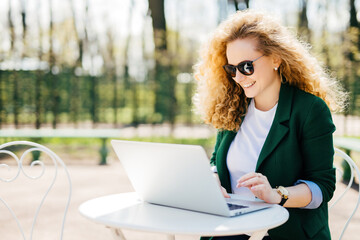 Curly-haired woman at park desk, happily messaging boyfriend on laptop. Pleasant reading, outdoor relaxation.