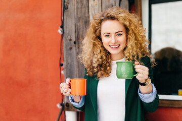 Blonde curly-haired woman with coffee mugs, happy expression, pleasing smile, ready to treat...