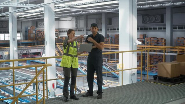 Caucasian Female Manager Talking to Multiethnic Male Distribution Specialist And Using Laptop Computer In Modern Warehouse Facility With Conveyor Belt. Products Being Packaged And Delivered To Clients
