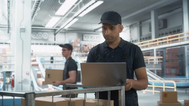Multiethnic Male Stocking Associate Doing Inventory On Laptop Computer, Checking Delivery Tracking Number On Boxes. Man Working In Warehouse Facility With Conveyor Belt Packing Products For Customers.