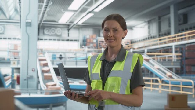 Modern Sorting Center: Female Stocking Associate Uses Laptop, Looking At Camera, Smiling. Transportation Logistics Warehouse Facility with Conveyor Loading Product Boxes To Deliver To Customers.