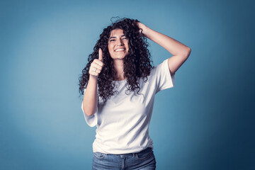 Naklejka premium Shot of happy confident curly woman with toothy smile, wears casual basic solid white t-shirt, expresses good emotions, enjoys nice day, doing thumb up symbol. Expressions
