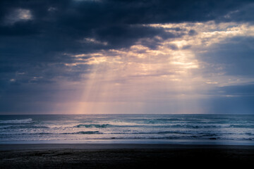 Sunset over Muriwai beach. Sun beams breaking through heavy clouds as waves splashing against the rocks. Auckland, New Zealand