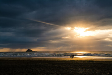 Sunset over Muriwai beach. Sun beams breaking through heavy clouds as a group of people stands by...