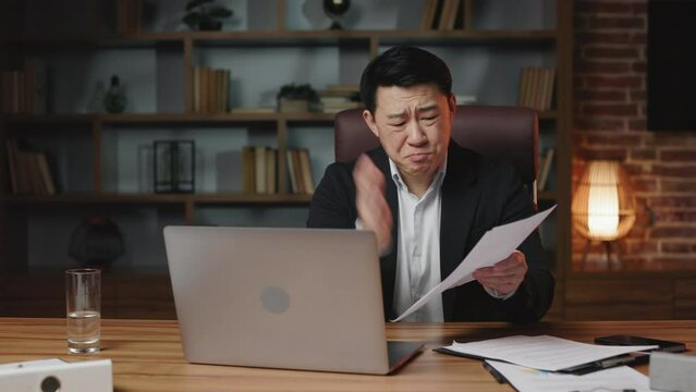 Angry male executive criticizing accountant's financial report during video call and tearing papers in front of computer screen in office. Strict asian professional feeling disappointed with bad work.