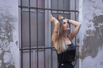 Fototapeta na wymiar Young woman, beautiful and blonde, with black tank top clinging to the grille of a window, with a sensual and flirtatious look. Concept sensuality, beauty, flirting.