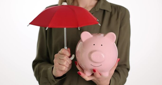 Woman holds pink piggy bank under red umbrella as insurance. Lady convinces to accumulate petty money. Good attitude towards finances