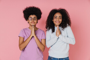 Two african women making pray gesture while standing isolated over pink background