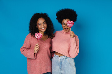 Two african women posing with lollipops while standing isolated over blue wall
