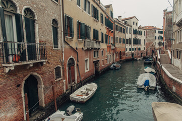 A picturesque Venice canal with boats lining the water, charming houses on one side, and a bridge with a sidewalk on the other.