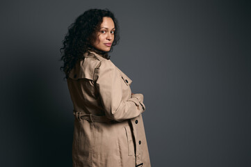 Beautiful Hispanic curly woman, wearing stylish beige trench, looking at camera through her shoulders, isolated on gray