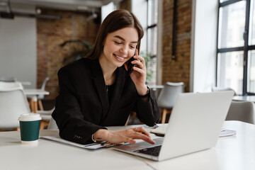Smiling business woman talking on mobile phone while working on laptop in office