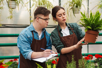 Beautiful woman florist helping man with down syndrome to handle with plants in greenhouse