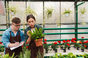 Man writing down notes in clipboard while woman florist teaching him to handle with plants in greenhouse