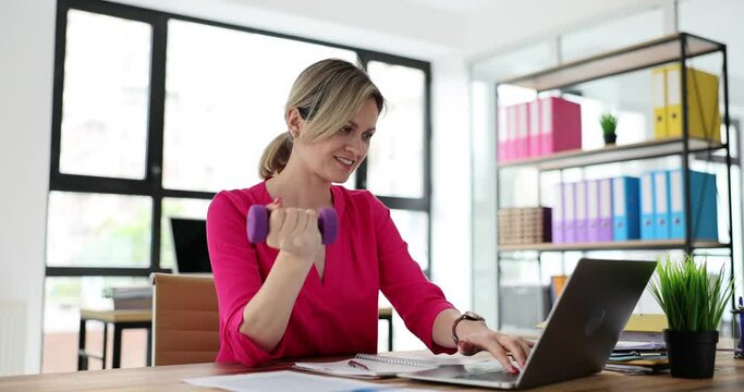 Young office manager does sports using purple dumbbell sitting at wooden table against laptop. Smiling lady does workout at work due to lack of free time
