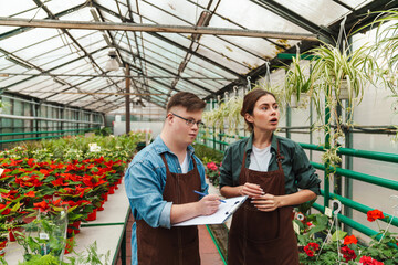 Man writing down notes in clipboard while woman florist teaching him to handle with plants in greenhouse