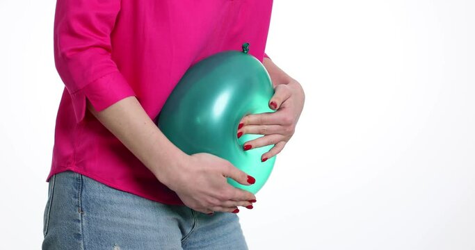 Female person in pink shirt compresses green balloon into body by pressing in middle. Lady tries to soothe pain at bloating and to burst balloon in light room