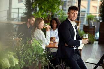 Focus on handsome businessman standing with beer glass and smiling. Blurred people on background. Team meeting. Concept of leisure time, business, communication, fun, alcohol drink