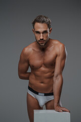 An attractive male model with a handsome physique and bare torso, posing in underwear while leaning on a gray crate, set against a gray background