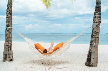 Young woman relaxing in wicker hammock on the sandy white beach on Mauritius coast and enjoying wide ocean view waves. Exotic countries vacation and mental health concept image.