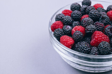 Close-up of glass bowl with red and black raspberries on gray background. Macro shot.