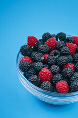 Close-up of glass bowl with red and black raspberries on blue background. Macro shot.