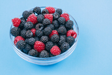 Close-up of glass bowl with red and black raspberries on blue background. Macro shot.