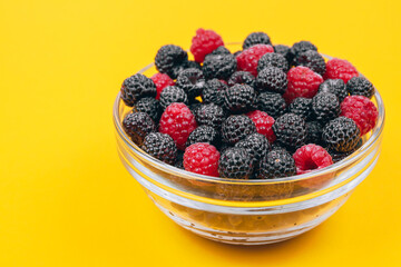 Close-up of glass bowl with red and black raspberries on yellow background. Macro shot.